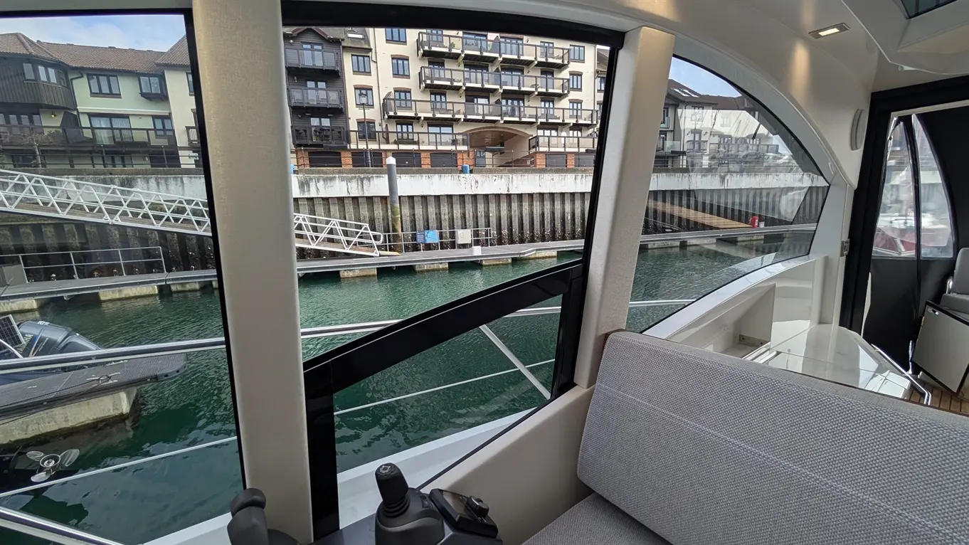 View from a boat interior showing a grey seating area, a marina with moored boats, and residential buildings in the background.